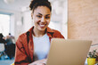 © BullRun - Portrait of happy female student smiling at camera during leisure time with laptop device in cafeteria, prosperous dark skinned hipster girl in casual wear sitting at table with new netbook