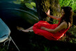 © Connect Images - Woman in red dress sitting under a tree by a pond touching the water surface with canoe nearby.