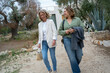 © Connect Images - Two women walking and laughing together outdoors near olive trees with a modern building in the background
