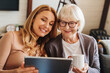© InsideCreativeHouse - Cheerful senior woman with cup of tea and her daughter using tablet at home