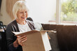 © InsideCreativeHouse - Happy senior grey-haired woman sitting on couch opening carton box received parcel package, internet order