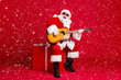 © deagreez - Full length photo of retired old man white beard hold guitar sit box play sing christmas carol song wear santa x-mas costume sunglass headwear leather boot isolated red color background