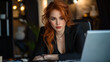 © Oksana - A woman with red hair in a business suit is focused on working at a laptop, sitting at a desk in a modern office