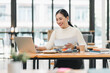 © kenchiro168 - young woman is working at desk in modern office, smiling as she reviews documents on her laptop. bright environment enhances her focused and positive demeanor.