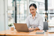 © kenchiro168 - young woman is working on laptop in modern office environment, showcasing focused and productive atmosphere. She is dressed in professional white shirt, with coffee cup and small plant on table,