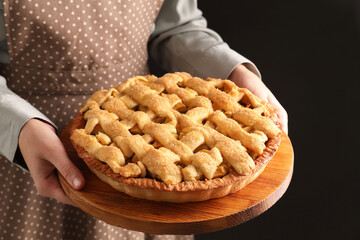 Sticker - Woman holding wooden board with tasty homemade apple pie on black background, closeup