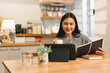 © kenchiro168 - woman is sitting at wooden table in cozy kitchen, smiling while holding cup and looking at tablet. She has notebook open in front of her, creating warm and inviting atmosphere.
