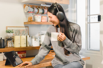 Wall Mural - Enjoying cozy moment in kitchen, woman with long hair wears headphones while holding cup and looking at tablet. warm atmosphere invites relaxation and focus.