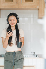 Wall Mural - Enjoying moment of relaxation, young woman with long hair listens to music on her smartphone while holding cup in modern kitchen. Her casual outfit and headphones create cozy atmosphere.