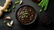 © fivan - A bowl of black bean sauce surrounded by garlic, ginger, scallions, and fermented black beans on a matte black background