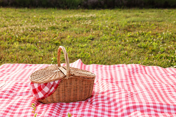 Wall Mural - Picnic wicker basket with napkin and red checkered blanket on green grass outdoors, space for text