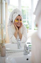 Woman in bathrobe with towel on her head smiles in the mirror, wearing golden under-eye patches. The scene highlights a refreshing skincare routine, beauty, and self-care in bright bathroom setting