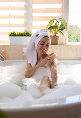 Wall Mural - A woman enjoys a relaxing bath in a bright, modern bathroom filled with natural light. Surrounded by plants and toiletries, she smiles while scrubbing her arm, creating a serene self-care moment.