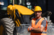 © zphoto83 - Construction worker in safety gear stands confidently near a yellow heavy machinery during daylight hours at a job site