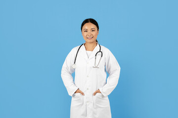 A healthcare professional Asian lady stands confidently in a white coat with a stethoscope around her neck. The bright blue background emphasizes her welcoming smile and dedication to patient care.