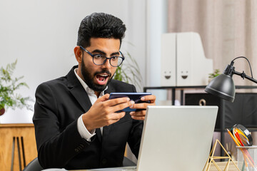 Indian businessman playing on smartphone mobile application celebrate winning racing or shooter game at office desk workplace. Professional manager freelancer man enjoy testing good work of new app