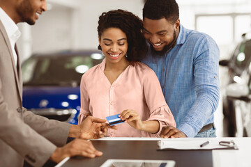 Buying Car Concept. Smiling Black Spouses Giving Credit Card To Automobile Seller In Auto Selling Showroom. Selective Focus