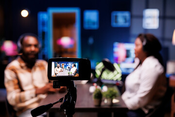 Wall Mural - Detailed view of professional camera used to film live broadcast between show presenter and man in home studio. Closeup of production equipment with host and guest chatting in blurry backdrop.