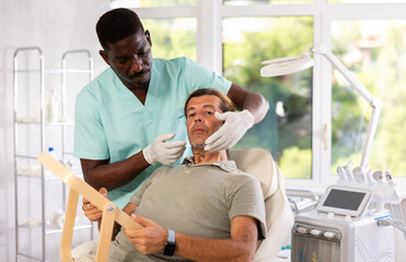 Adult man looking in mirror with interest while skilled African American male cosmetologist showing results of hardware facial procedure in office of aesthetic medicine clinic