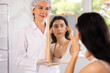 © JackF - Young female client examining her face in mirror after cosmetic procedure in clinic