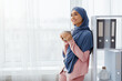 © Prostock-studio - Coffee Break. Cheerful Arabic Businesswoman In Hijab With Cup Of Hot Drink Standing Near Window In Office, Empty Space