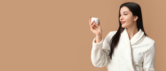 Poster - Beautiful young woman in bathrobe holding jar of cream on beige background with space for text