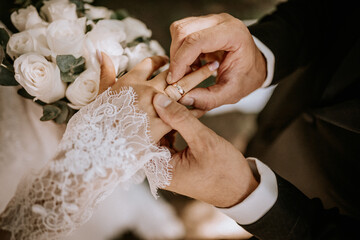 A close-up of a groom placing a wedding ring on the bride’s finger during the ceremony, with her lace sleeve and white rose bouquet visible.