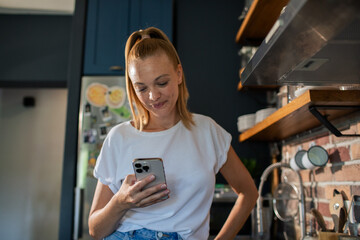 Wall Mural - Smiling young woman using smartphone in modern kitchen