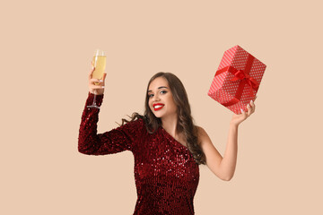 Sticker - Young woman with gift and glass of champagne on beige background. New Year celebration