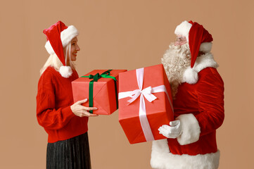 Poster - Santa Claus and beautiful mature woman in Christmas hats with gift boxes on brown background