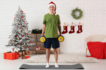 Poster - Young happy man in Santa hat training with dumbbells in living room decorated for Christmas