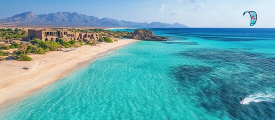 An aerial view of a beautiful beach with turquoise water and a kite surfer in the distance. The beach is surrounded by mountains and vegetation.