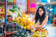 © Itza - Young Latina woman happily picking mangoes at a vibrant market in Mexico City, Mexico.