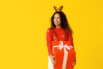 Wall Mural - Happy young African-American woman in Christmas sweater and deer horns with gift box on yellow background