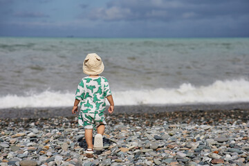 A toddler in a cute outfit and a sunhat walks along a rocky beach near the ocean as waves crash nearby. The scene captures the child's adventurous spirit and the natural beauty of the stormy 