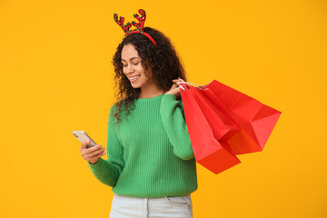 Beautiful young African-American woman in reindeer horns with mobile phone and shopping bags on yellow background. Christmas celebration