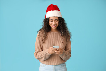 Canvas Print - Beautiful young African-American woman in Santa hat with mobile phone on blue background. Christmas celebration