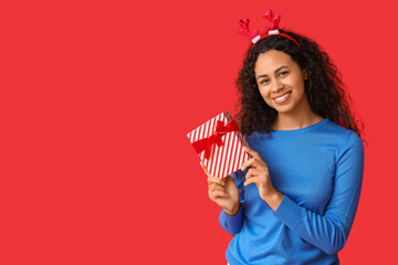 Sticker - Beautiful young African-American woman in reindeer horns with Christmas gift box on red background