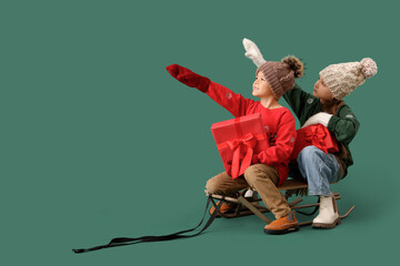 Poster - Cute happy little children with Christmas gift boxes sitting on sledge and pointing at something against green background