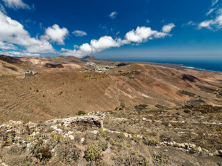 The volcanic landscape of Lanzarote features barren, rugged terrain with layered hills and sparse vegetation. The distant ocean contrasts with the dry earth under a clear blue sky scattered with cloud