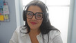 © Krakenimages.com - Young woman working indoors wearing glasses and headphones in an office setting with a white shirt and long dark hair, looking confident and focused