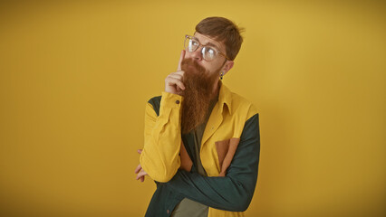 Wall Mural - A thoughtful young man with a beard and glasses posing against a yellow background, exuding a hipster vibe.