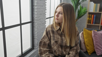 Poster - A thoughtful young blonde woman in a plaid shirt sits indoors near a window, looking away in a modern living room.