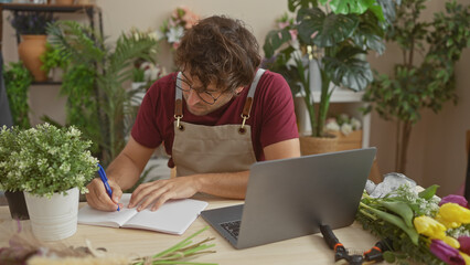 Poster - A focused hispanic man with a beard writes in a notebook at a flower shop, surrounded by plants and a laptop.