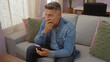© Krakenimages.com - Man sitting in living room on sofa in home holding smartphone with thoughtful expression and hand on chin