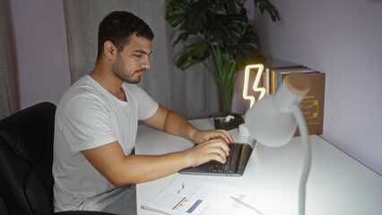 Young man working on his laptop at home, sitting in a neat and modern living room with documents scattered on the desk, illuminated by a stylish lamp in a serene indoor setting.