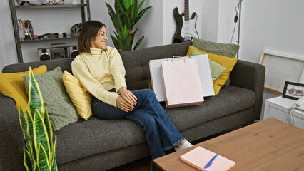 Wall Mural - Smiling young woman sits on couch in a modern living room with shopping bags, feeling relaxed and content.