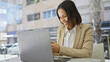© Krakenimages.com - A professional hispanic woman enjoys working on her laptop, set against a blurred cityscape background.