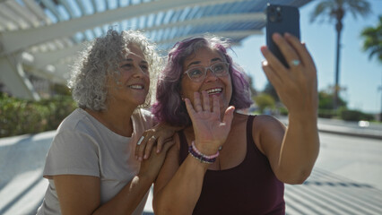 Poster - Two mature women with curly hairstyles are smiling and waving while taking a selfie outdoors in a modern urban area