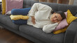© Krakenimages.com - A mature hispanic woman in pain lies uncomfortably on a sofa in a home setting, clutching her stomach.